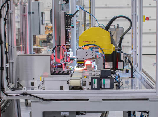 An automated industrial robot arm, identified as "FANUC LR Mate 200iA," performs a task within a protective enclosure. Red light shines from the robot's arm over the work area.