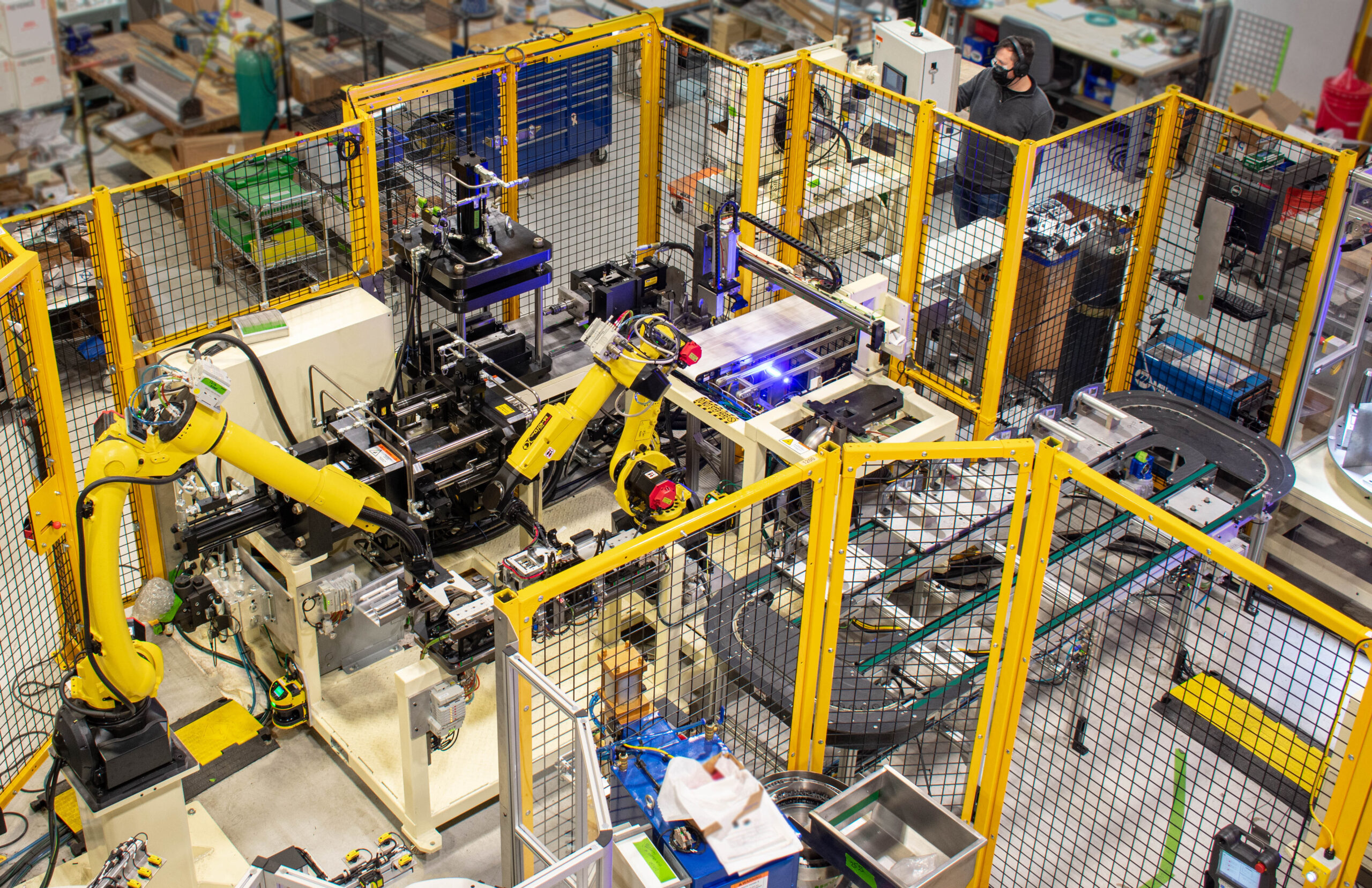 High-angle view of a brightly lit factory floor featuring a complex automated assembly line with several yellow robotic arms working on various components. The machinery is enclosed by yellow safety barriers.