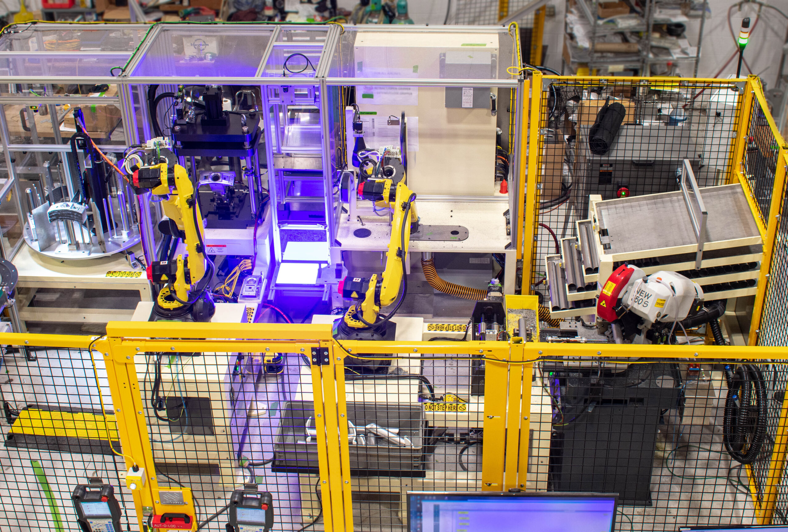 An overhead view of an automated manufacturing cell with two bright yellow robotic arms enclosed within clear plastic housings and yellow safety guarding.
