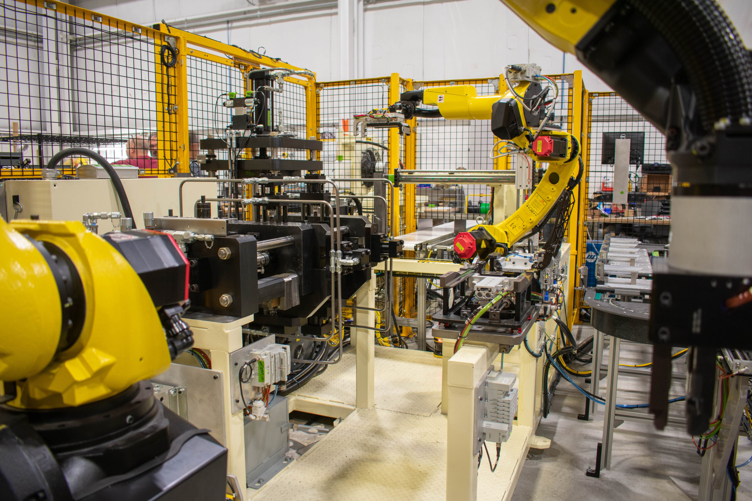 Automated production line with yellow robotic arms performing manufacturing tasks within a fenced enclosure.