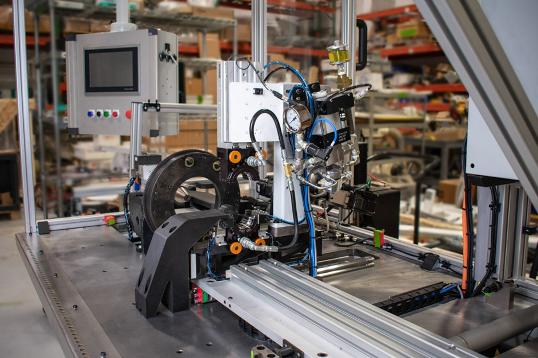 A semi-automated manufacturing machine sits on a metal table in a warehouse setting. The machine features a central control panel with a screen and a series of colored buttons.