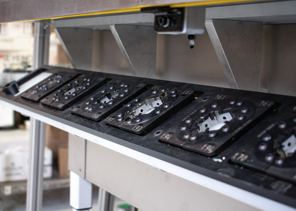 A close-up of a metal press machine shows a shelf holding several black, square die plates. Each plate has a circular opening in the center surrounded by screw holes.