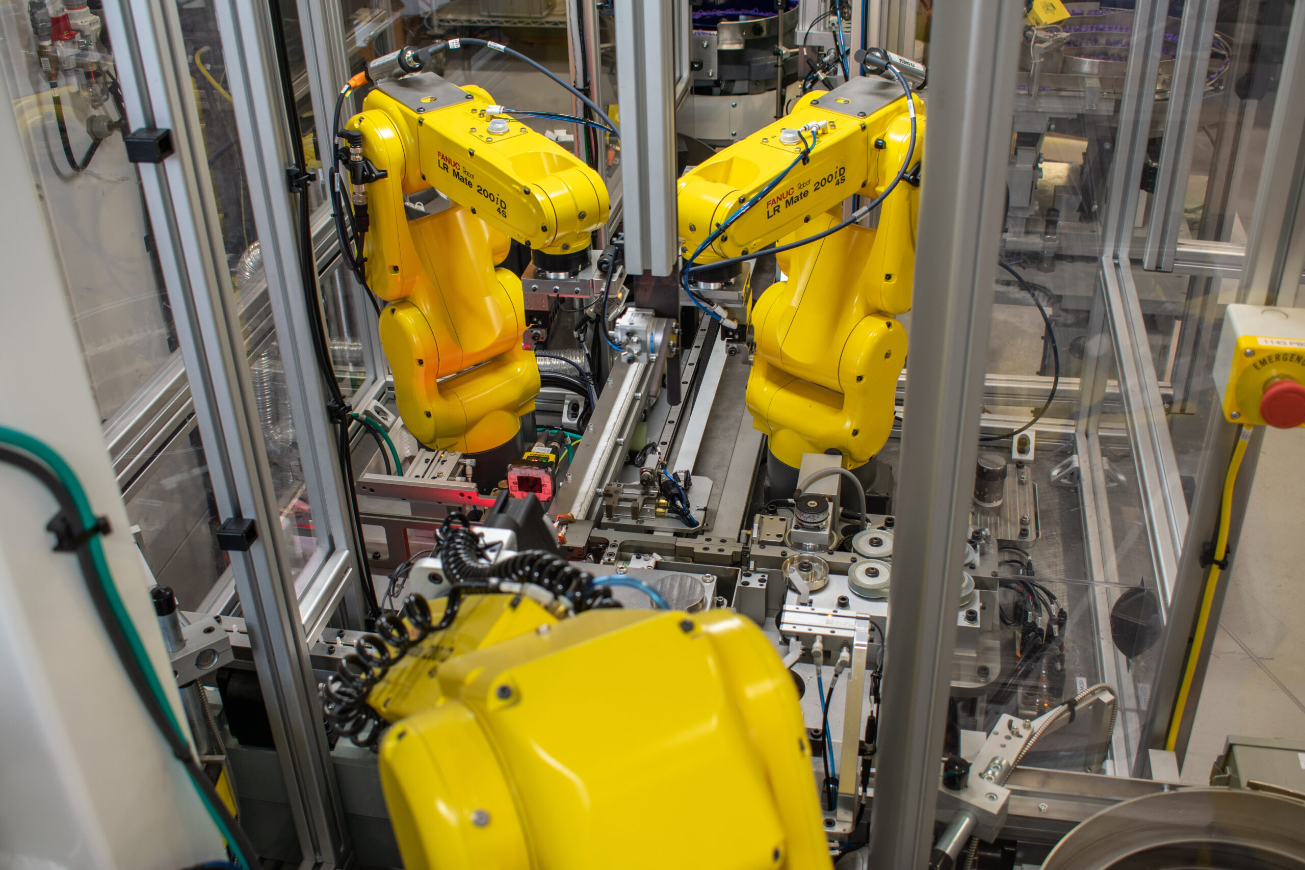 Image shows three yellow industrial robot arms positioned within a metal and glass-enclosed automated assembly process