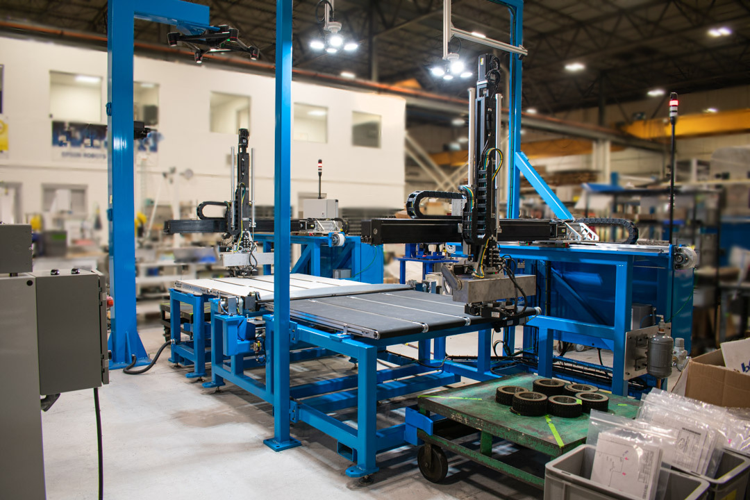 Assembly line features blue metal frames supporting multiple conveyor belts and robotic arms. Components are positioned on the belts ready for processing.