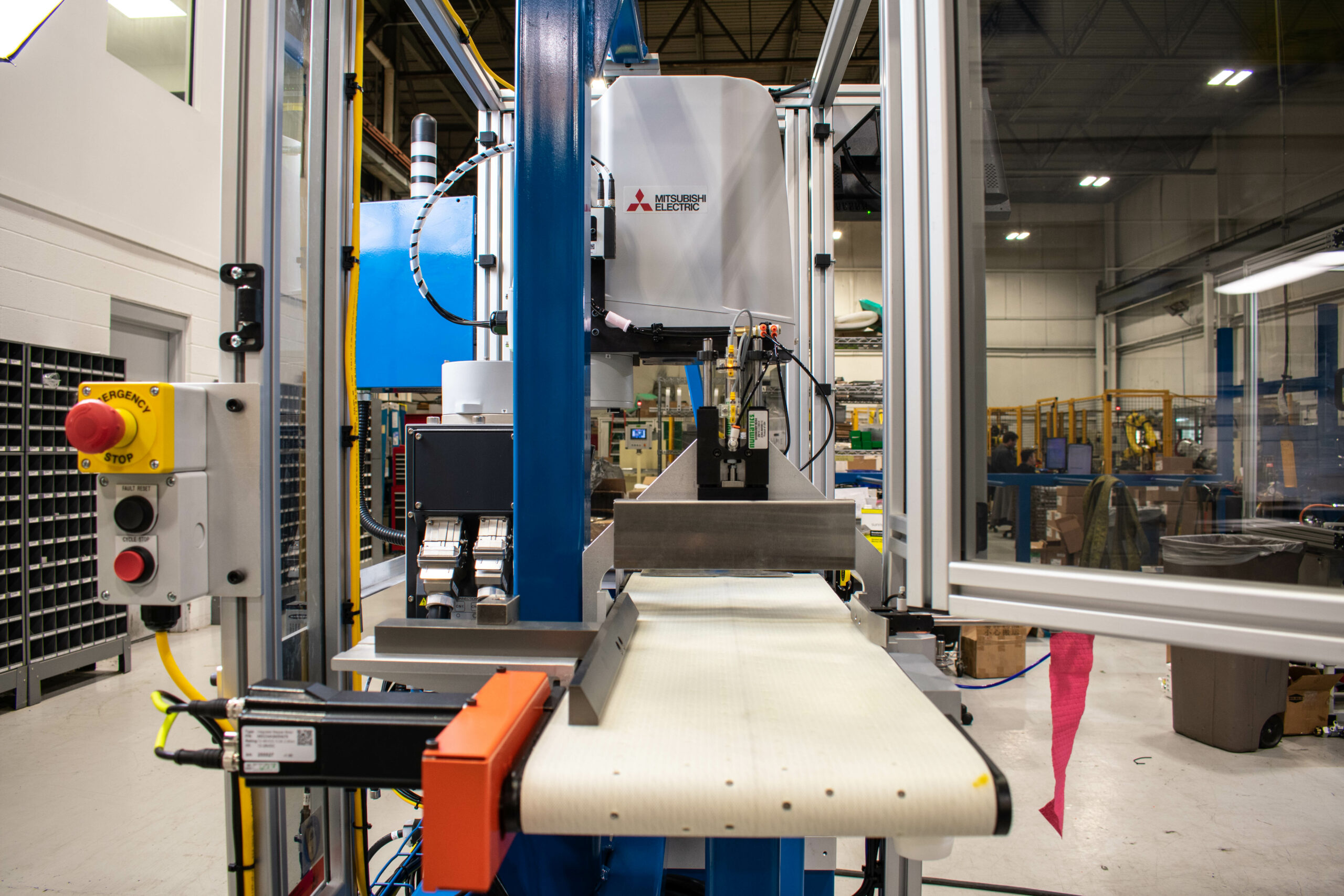 Industrial automation machine with a conveyor belt, Mitsubishi Electric robotic system, emergency stop button, and safety enclosure in a factory setting.