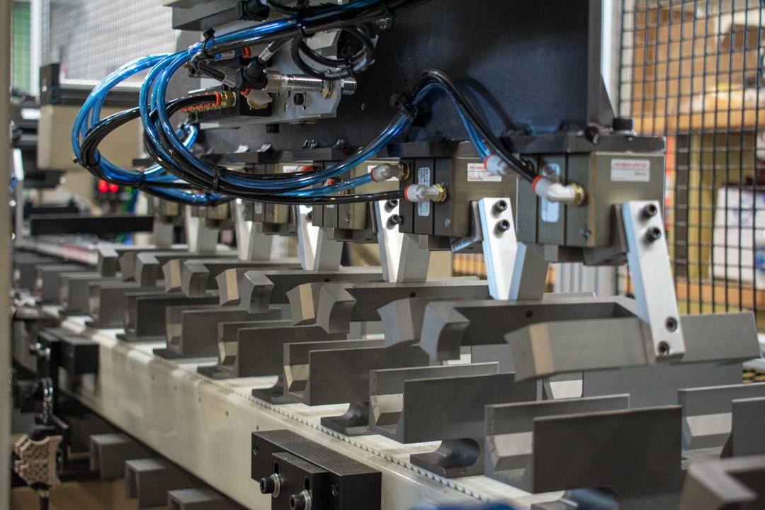 Close-up of automated industrial machinery featuring a series of metal clamps aligned over a conveyor. The clamps have pneumatic connections, and blue tubing provides air power.