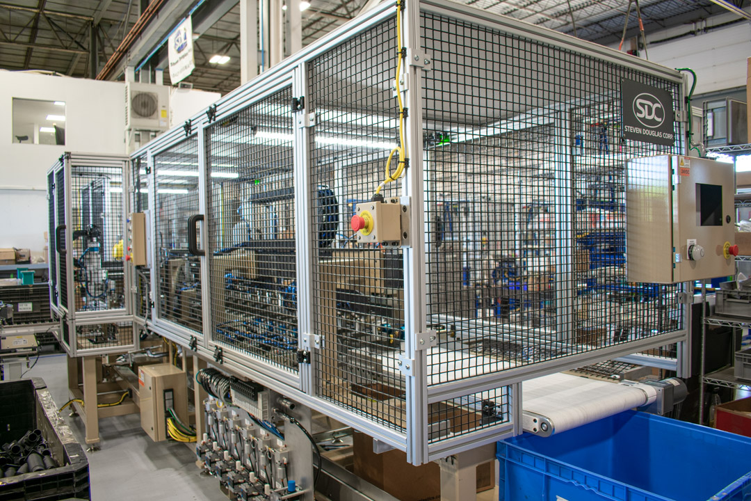 A large, automated industrial machine is enclosed by a silver aluminum frame covered in a black wire-mesh cage for safety.
