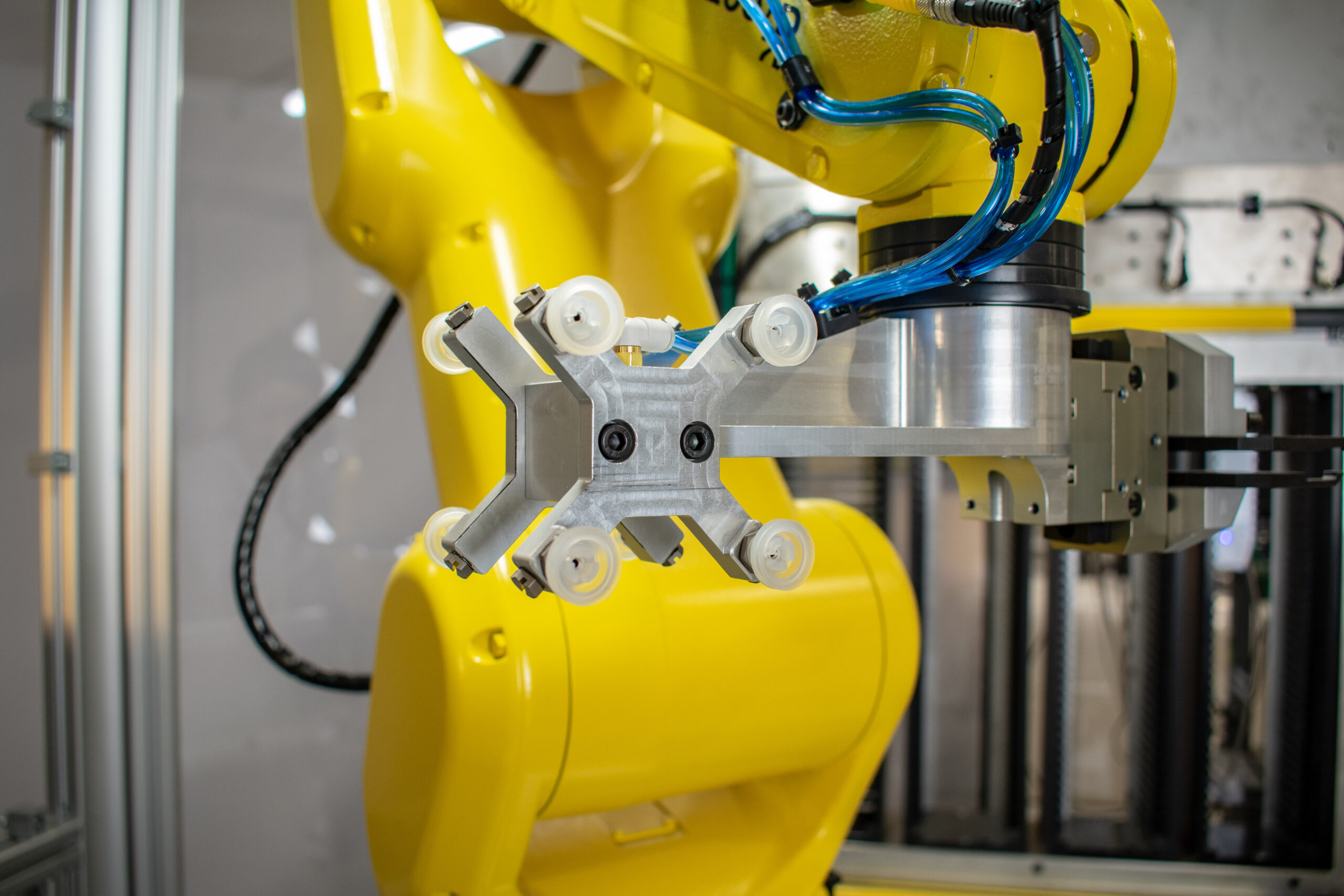 Close-up of a bright yellow industrial robot arm, focusing on its tool head. The tool head is a silver and grey apparatus with small, clear rollers. Blue tubing and black cables connect to the arm.