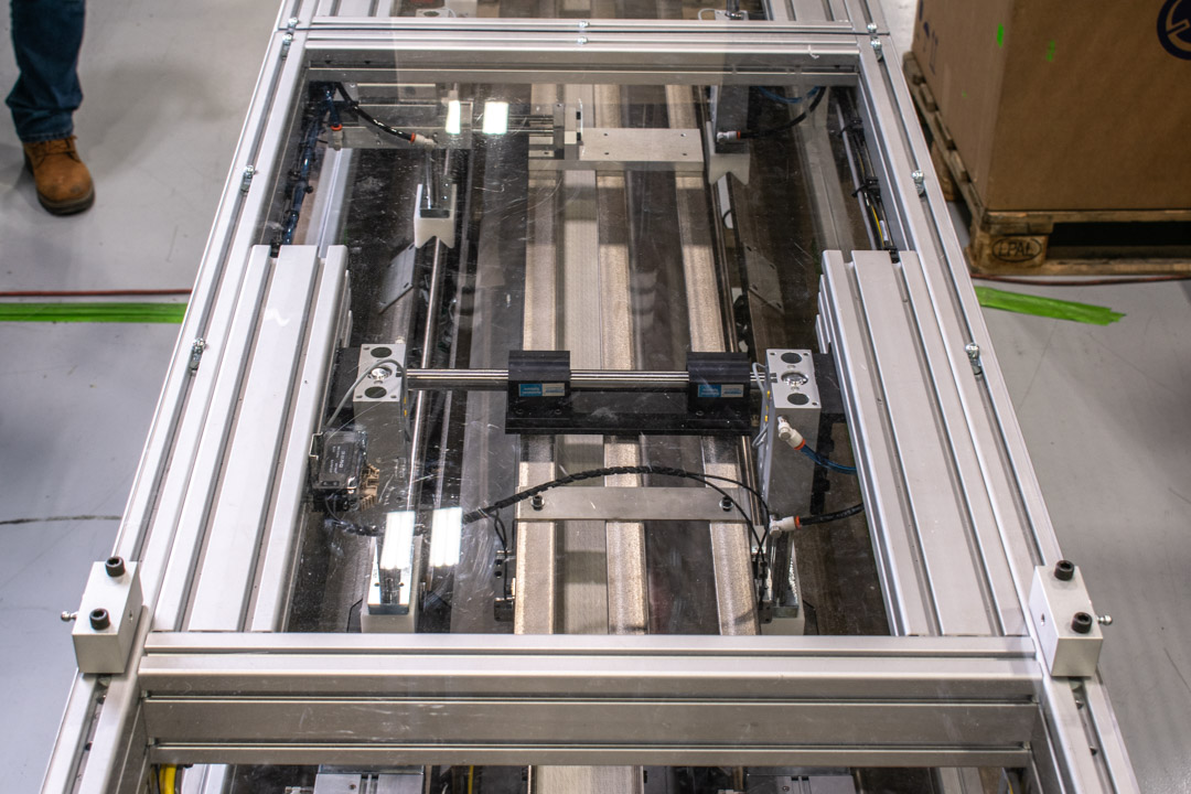 Overhead view of an automated machine with a clear safety enclosure. The machine has linear actuators mounted on an aluminum frame with multiple parallel rails.