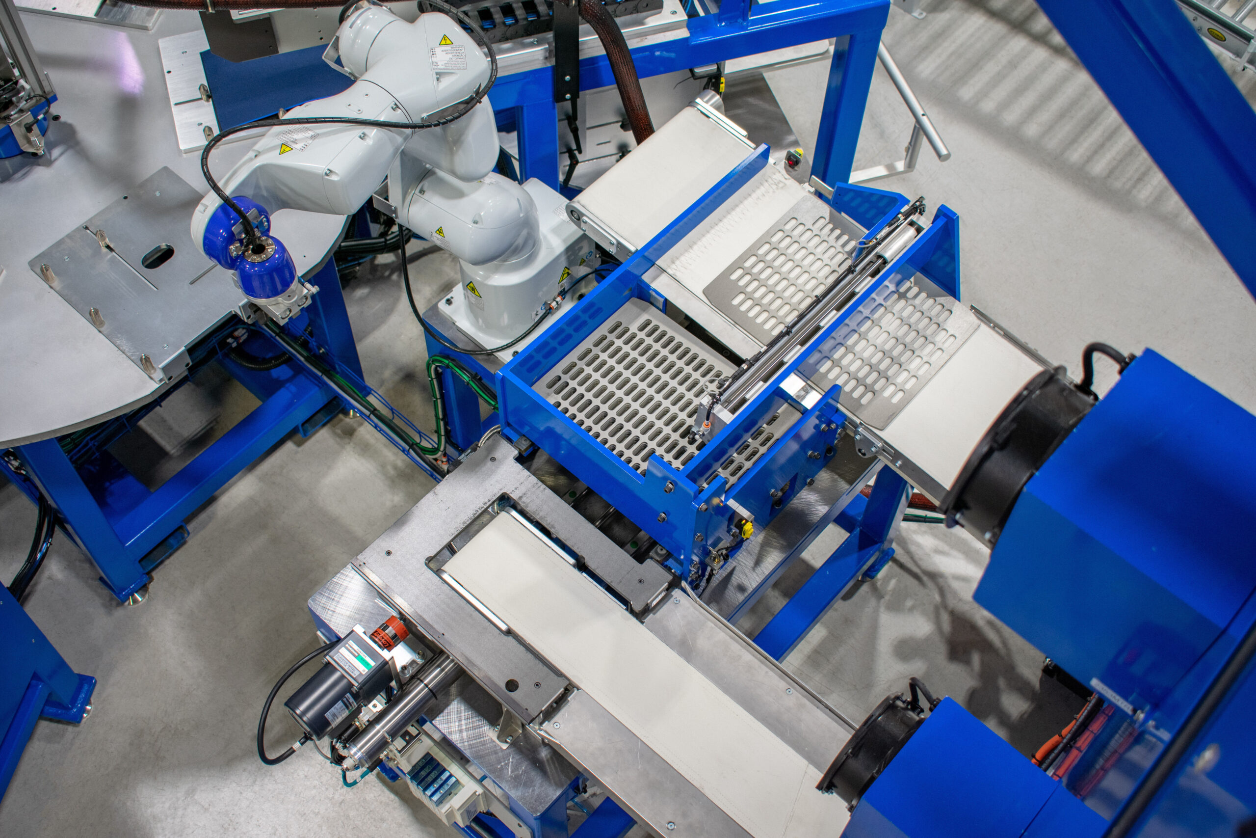 A white robotic arm with a blue attachment sits next to a circular metal platform. Blue machinery with conveyor belts and trays with uniform grids moves materials along the line.