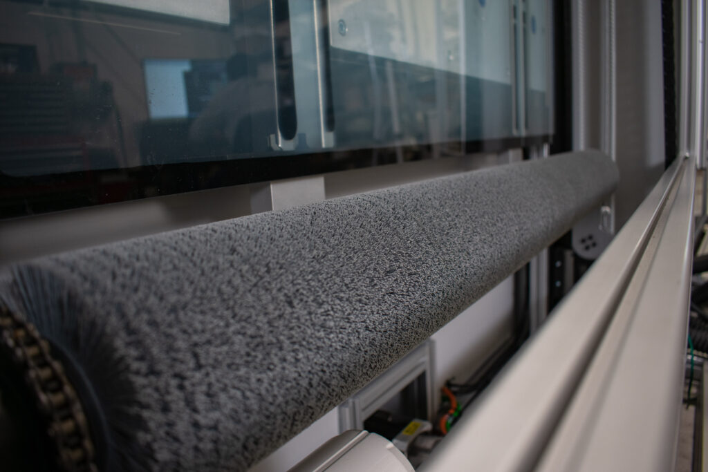 Large, uniformly gray-colored roller set within an industrial machine. Above the roller are clear glass panels, and the metal frame of the machine is visible, offering a glimpse into the inner workings.
