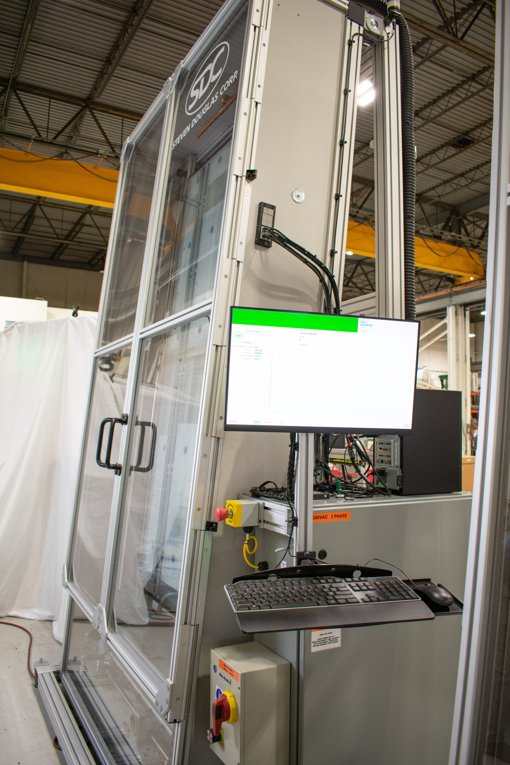 Glass-enclosed testing area with large handles. A computer workstation is attached to the machine, showing graphical data on its monitor. Emergency stop buttons and control panels are visible.