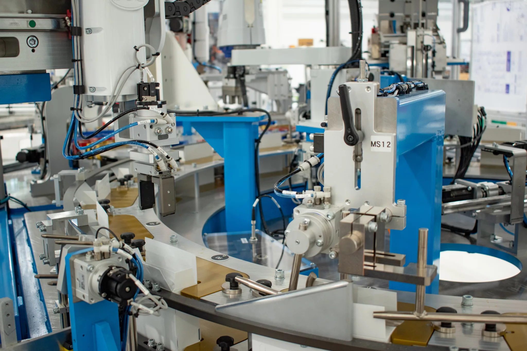 The scene features robotic arms with precision tools poised over a circular conveyor system. Various actuators, valves, and tubing are visible.