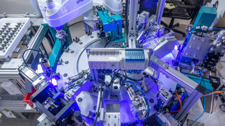 A complex, overhead shot shows a highly automated industrial assembly line. Robots manipulate parts around a central circular platform, utilizing blue lights for enhanced visibility.
