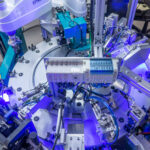 A complex, overhead shot shows a highly automated industrial assembly line. Robots manipulate parts around a central circular platform, utilizing blue lights for enhanced visibility.