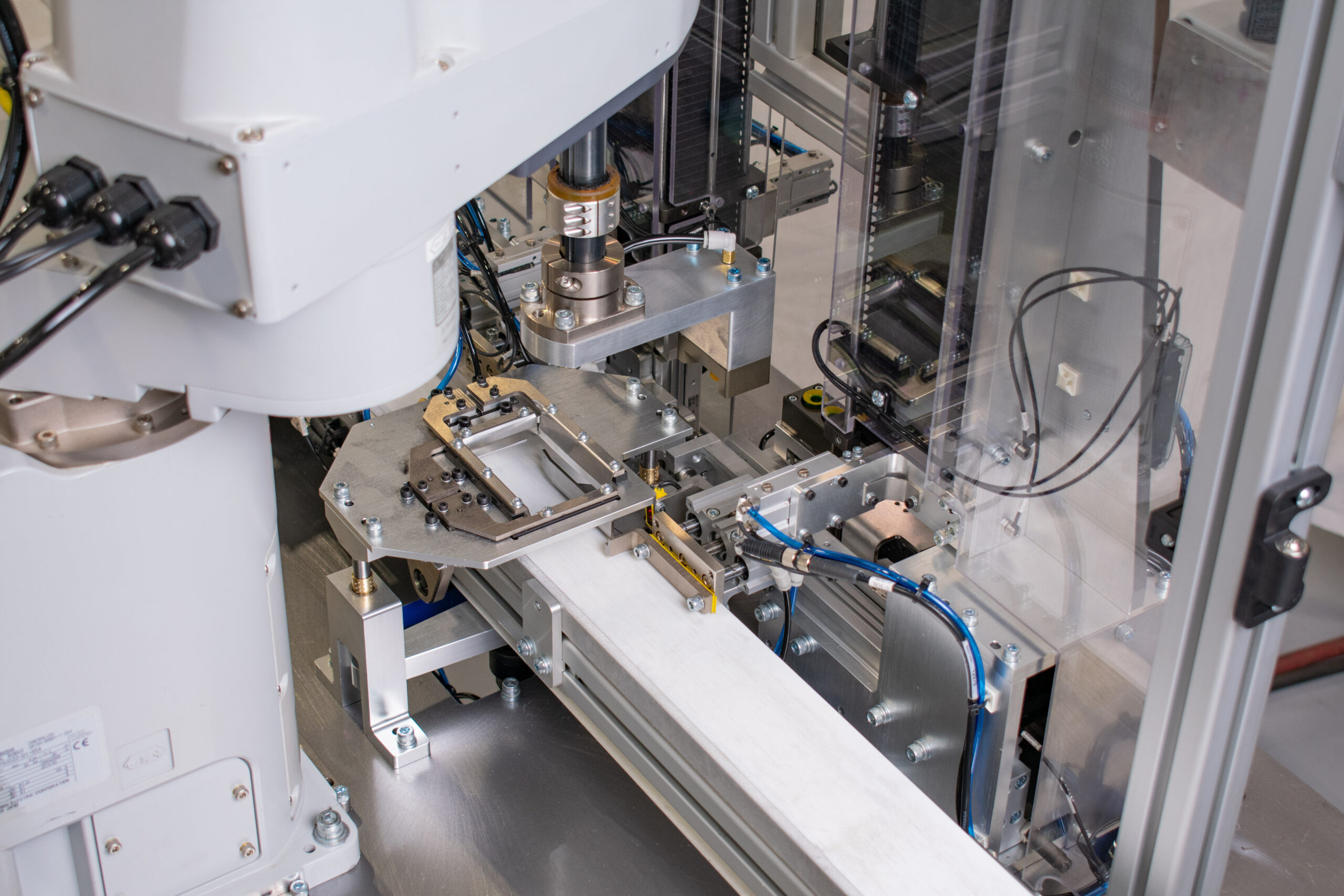 Robotic arm appears to be in the process of manipulating a rectangular component held within a metal fixture on a conveyor belt. The machine is constructed from metal framework with intricate mechanisms, gears, and clear safety shields visible.