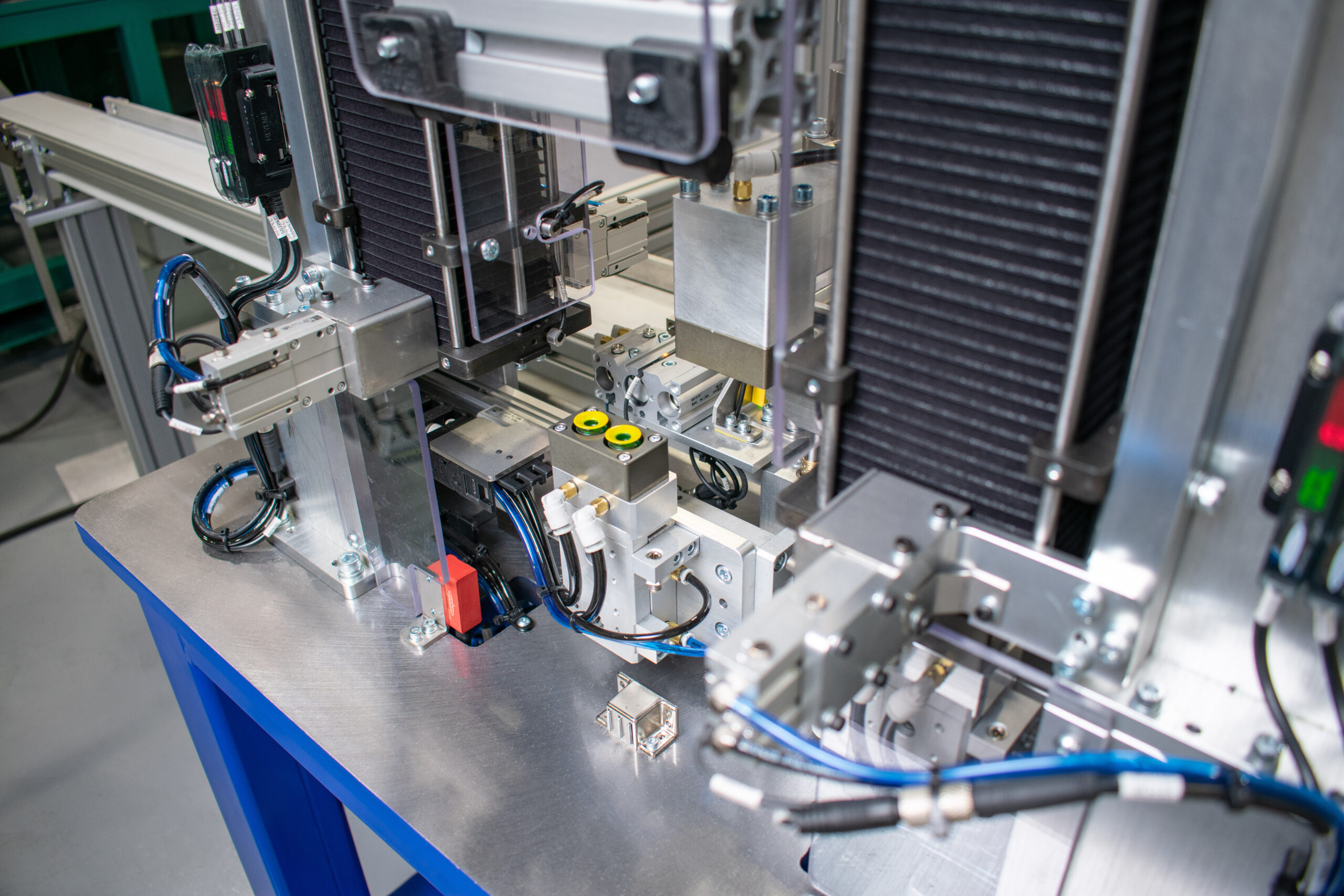 Close-up of an automated assembly machine composed of metal components on a blue table, with a brushed metal surface. The machine features numerous pneumatic actuators, sensors, and wires.