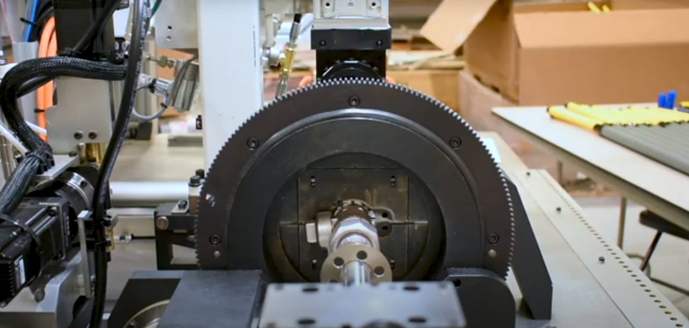 Close-up of an industrial machine processing a metal pipe. The machine features a circular, toothed gear surrounding the pipe, with various cables, hoses, and mechanical components visible.