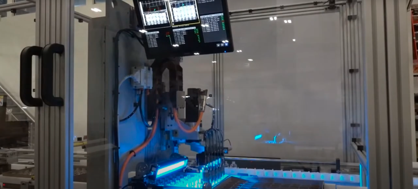 Automated laboratory machine inside a transparent enclosure. The machinery consists of a robotic arm with multiple nozzles positioned over a tray filled with small vials.