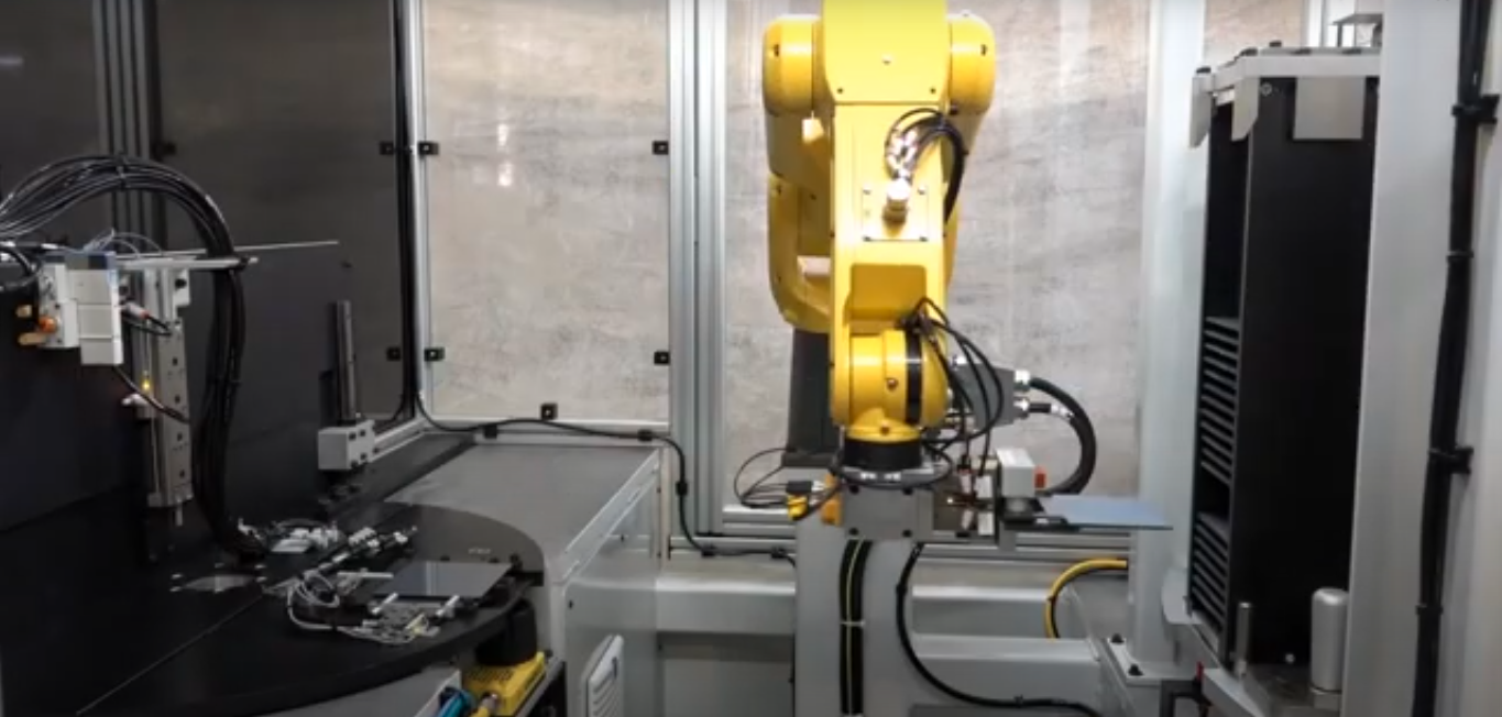A FANUC yellow robot connected to multiple wires operates machinery in a factory setting, showcasing advanced automation in robotic material handling.