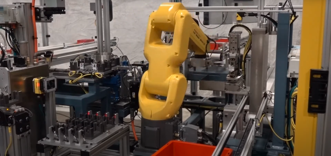 A yellow industrial robotic arm interacting with components on an automated assembly line. Multiple workstations and sensors are visible, indicating a complex manufacturing process.