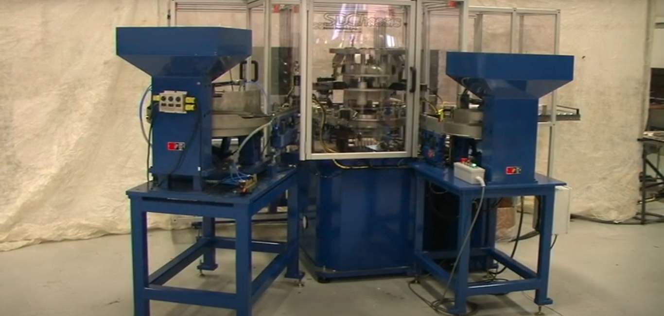 Industrial palletizing machine with a blue structure and clear machine guarding. Multiple wires and cords are running from the machine. In front of a tan curtain backdrop.