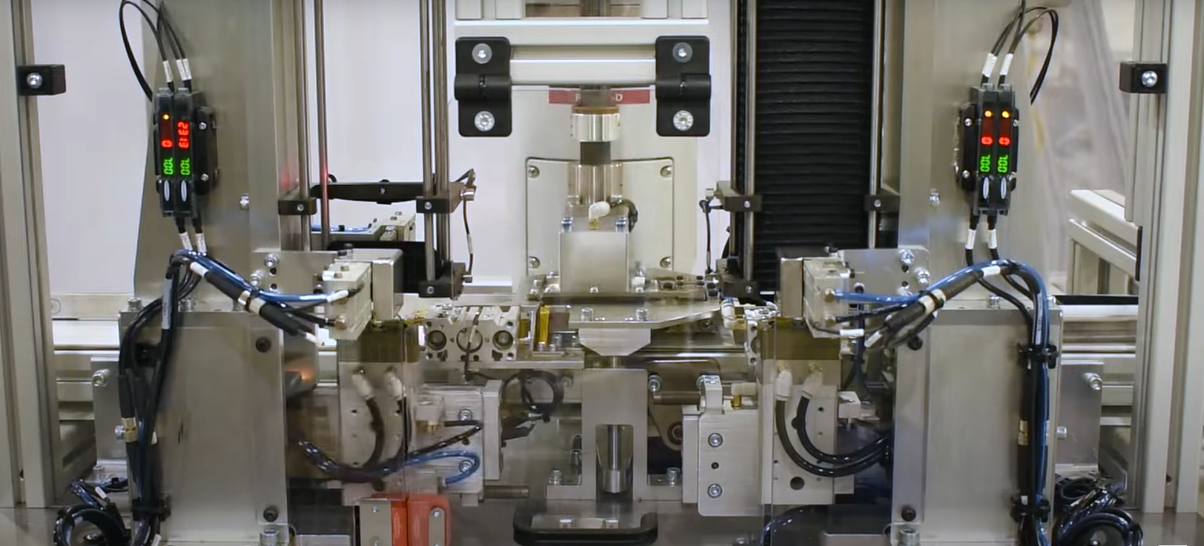 The machinery consists of silver metal elements, including several visible gauges with digital numerical displays which read "100", pneumatic cylinders, and intricately routed blue and black wiring.