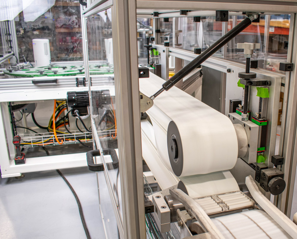 Large roll of white material is being fed through machinery, moving along a conveyor belt. Features a metal frame, clear protective shields, and various cylinders and sensors.