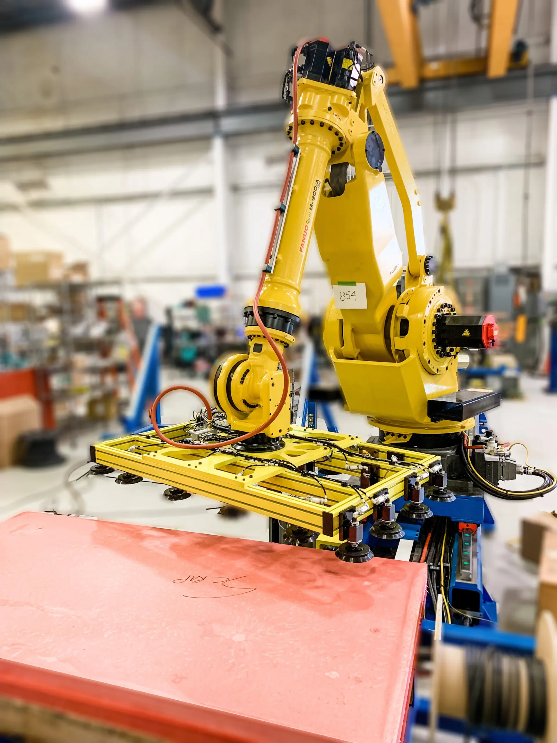 A yellow industrial robot arm in a bright factory setting extends towards a large, flat rectangular material. The robot's arm is equipped with a square yellow platform containing multiple suction cups, these affix to the material.