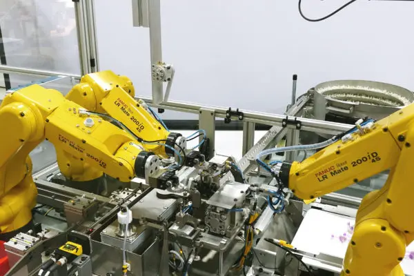 Three yellow FANUC LR Mate 200iD robot arms working together on one single gray metallic assembly with machine guarding in the background.