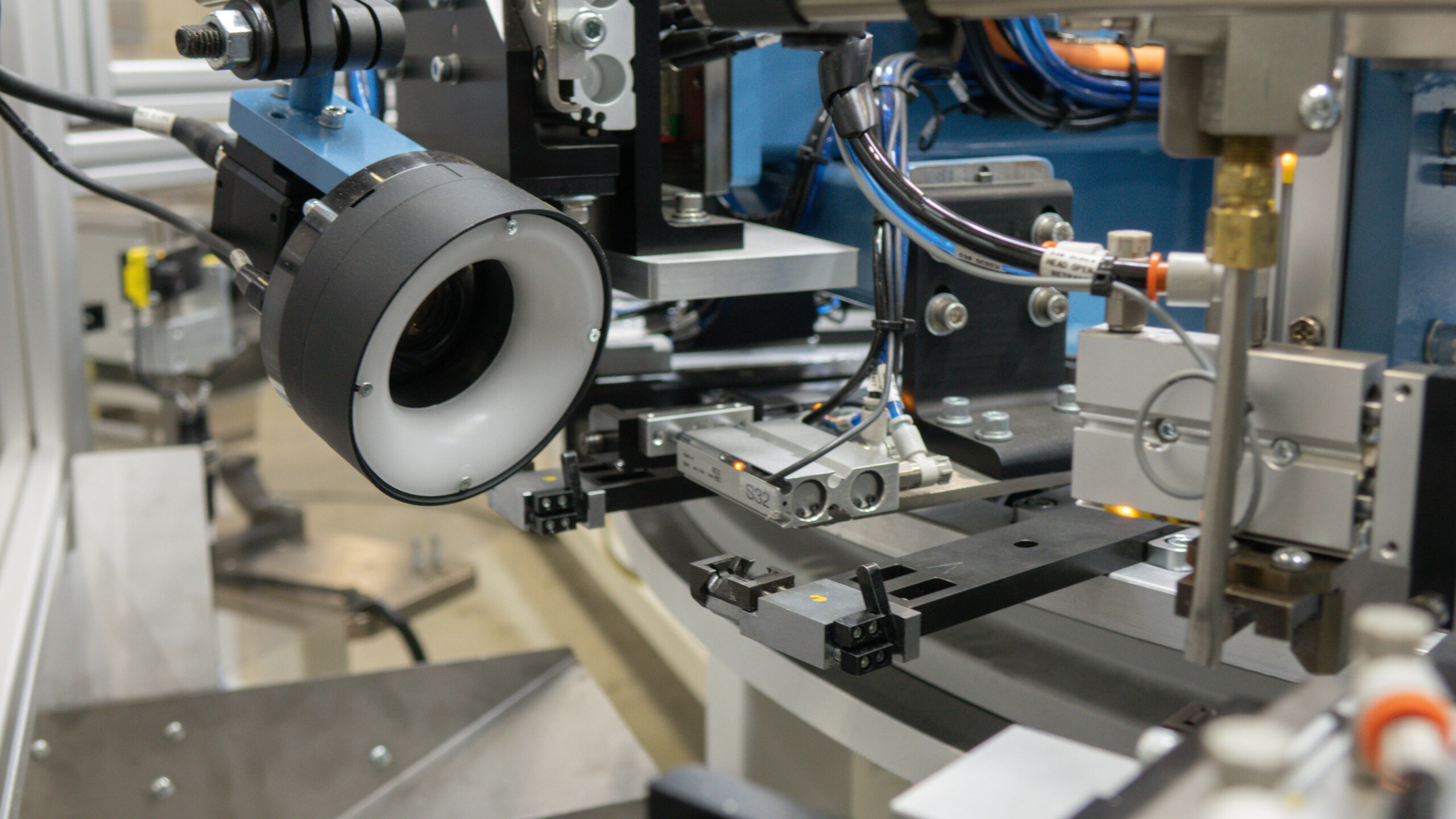Close-up of an automated industrial machine, showing a camera with a ring light, robotic arm, and pneumatic controls in a factory setting.