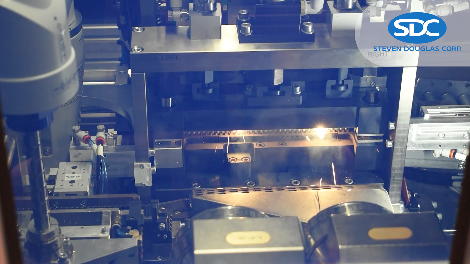 Industrial laser cutter in operation. Bright sparks are visible as the laser beam cuts through a metal component. Parts of the cutting machine are highly reflective, and a logo for (SDC) is visible.