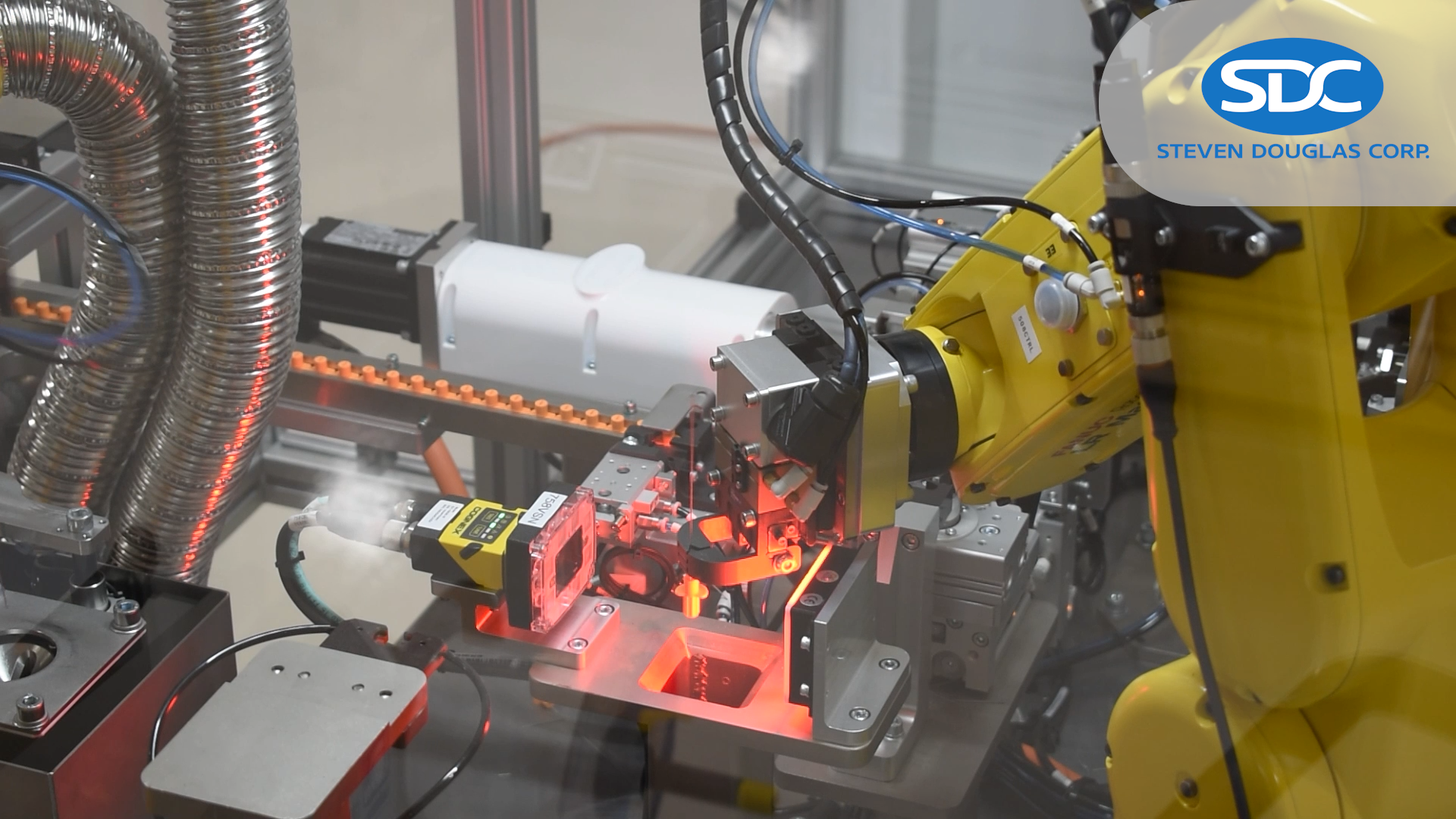A robotic arm, branded with the SDC logo, is shown manipulating components in an automated process. Robot is yellow, and appears to be placing or inspecting an item that is illuminated by a red light.