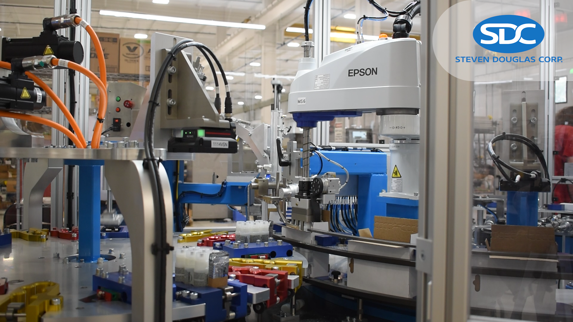 Automated robotic assembly line in a manufacturing facility, with blue and white robotic arms assembling small colored components. The Steven Douglas Corp logo is overlaid.