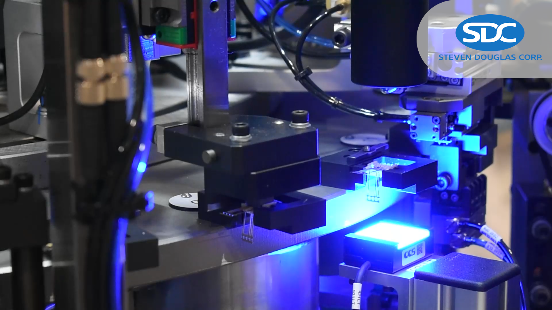 Automated assembly equipment by Steven Douglas Corp (SDC) working on small electronic components. The machinery features a robotic arm and blue LED lighting.