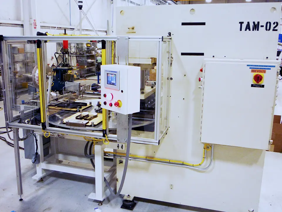 Machine is white and contains safety guarding around a rotating table. Control panel with a digital display and multiple red and white control buttons. A high voltage warning label is prominent on the control box.