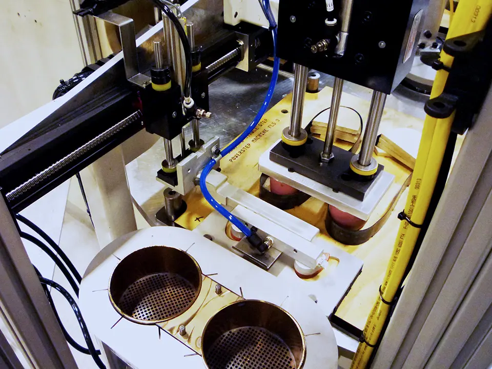 High angle shot of an industrial automation apparatus; vertical composition focused on tooling, precision components, and pneumatic cylinders above two circular openings on a white work surface.