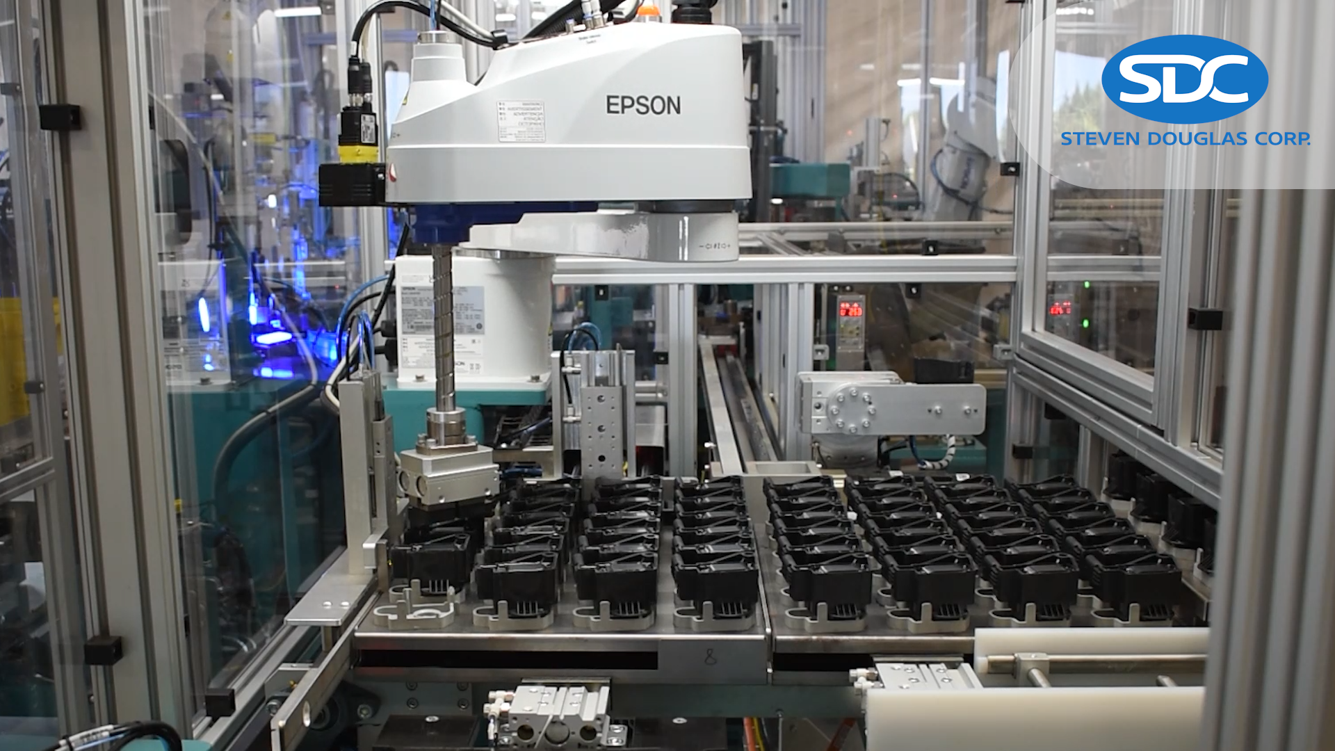 Automated assembly machine with multiple pneumatic cylinders visible in the foreground. The SDC (Steven Douglas Corp) company logo is in the upper-right corner.