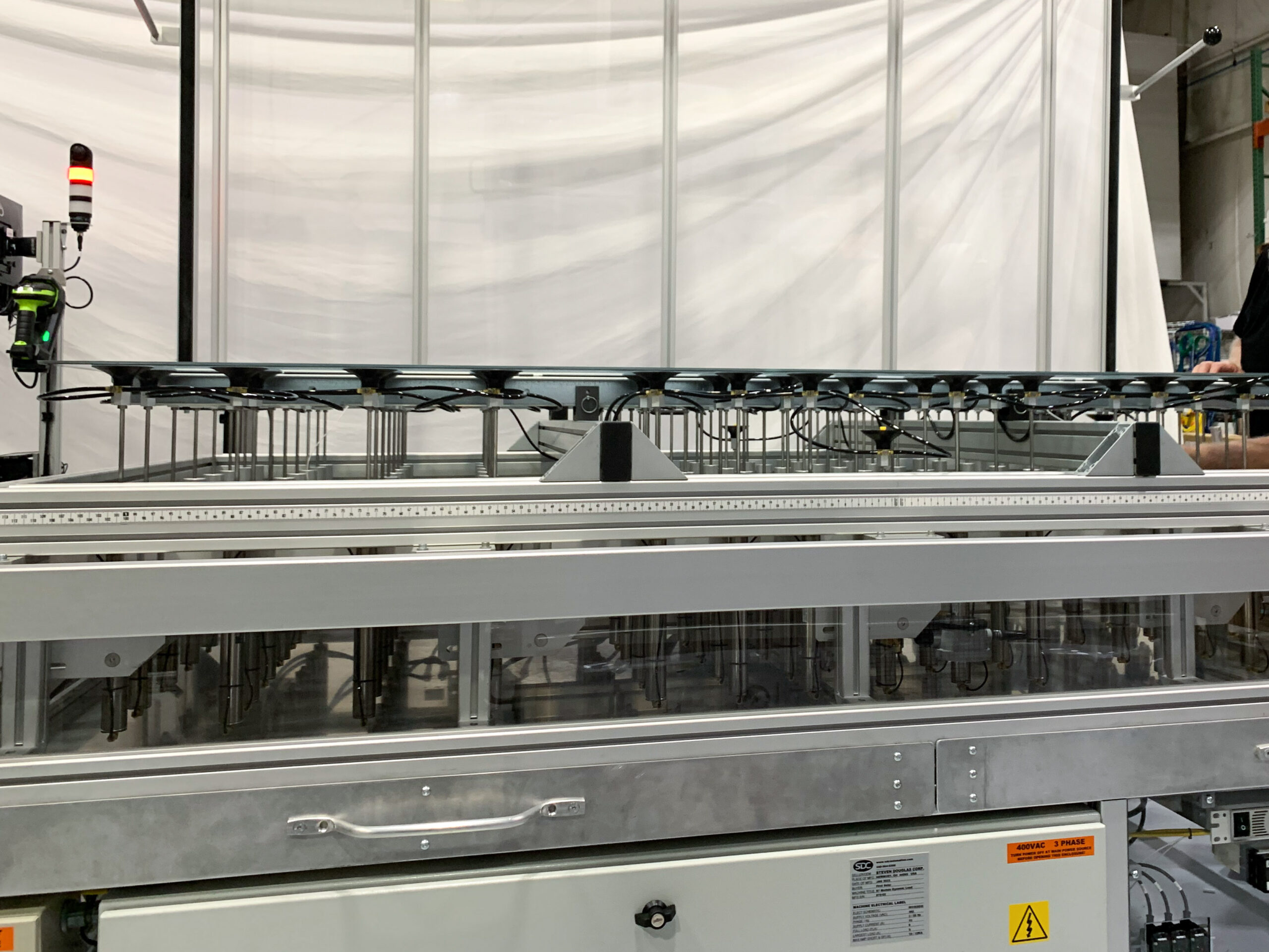 Close-up of a production line machine in a factory setting. Clear glass panels are held in place by an array of black suction cups attached to metal rods within a horizontal frame.