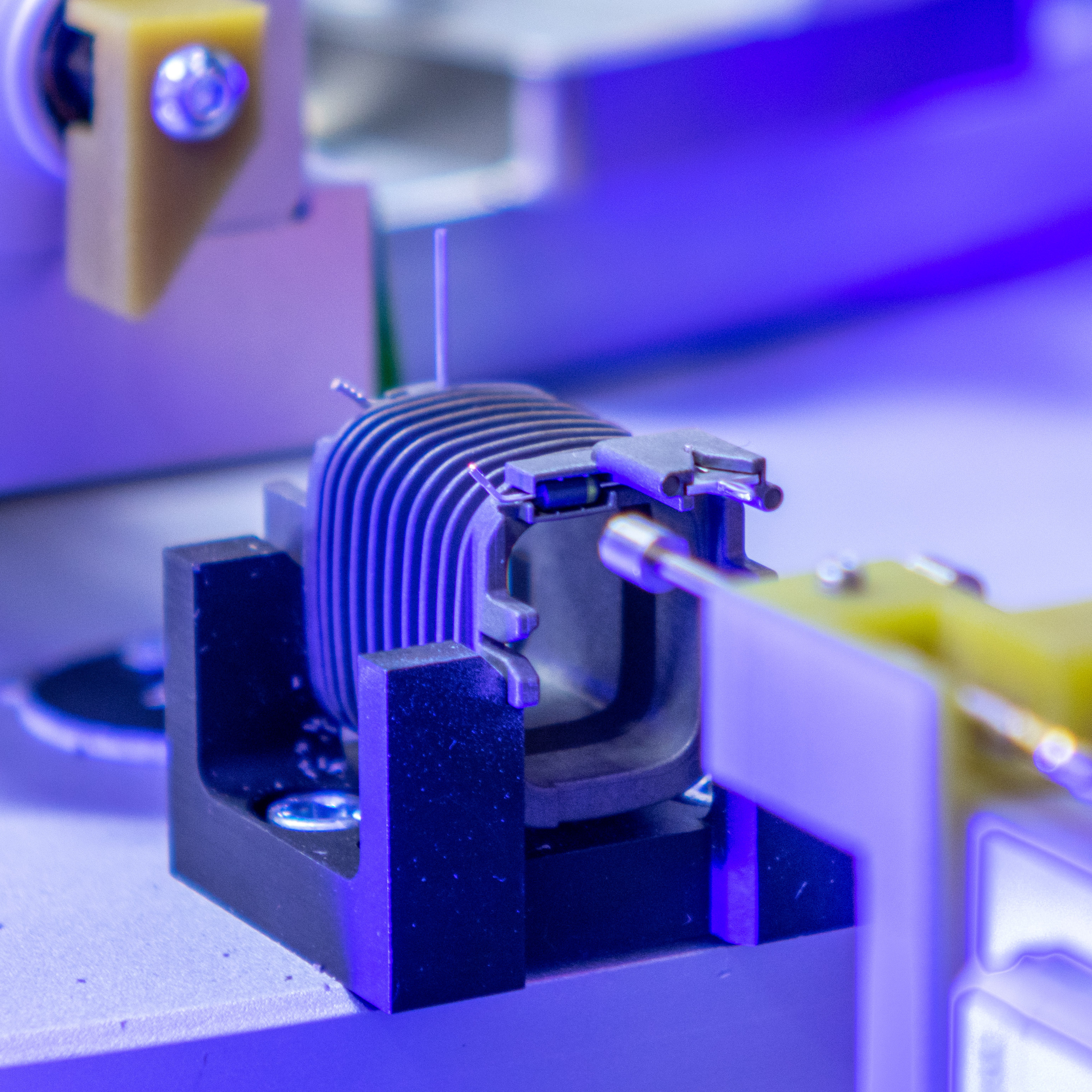 A close-up of an electromagnetic coil being processed in a factory. The coil is held by a black bracket on a light gray surface. The image is bathed in a blue light.