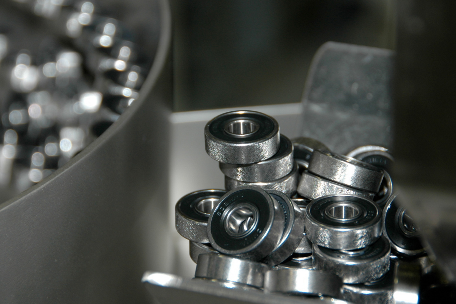 Close-up shot of a pile of metallic ball bearings, each featuring a silver ring surrounding a dark grey center. The bearings are stacked imperfectly on a gray metal surface.