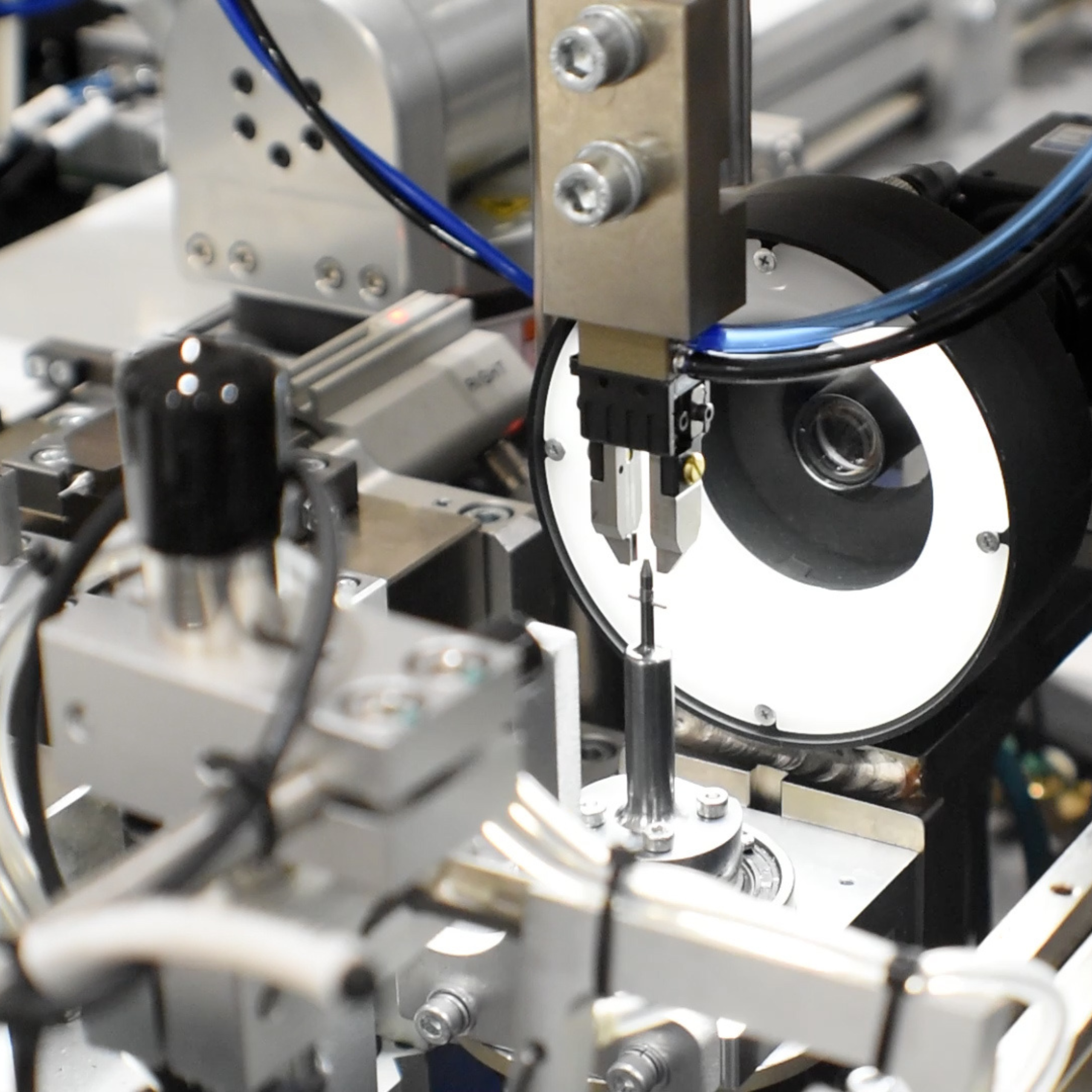 Machine is made up of silver metal components of clamps, arms, and sensors. A bright, round backlight illuminates the work area where a process is being performed.