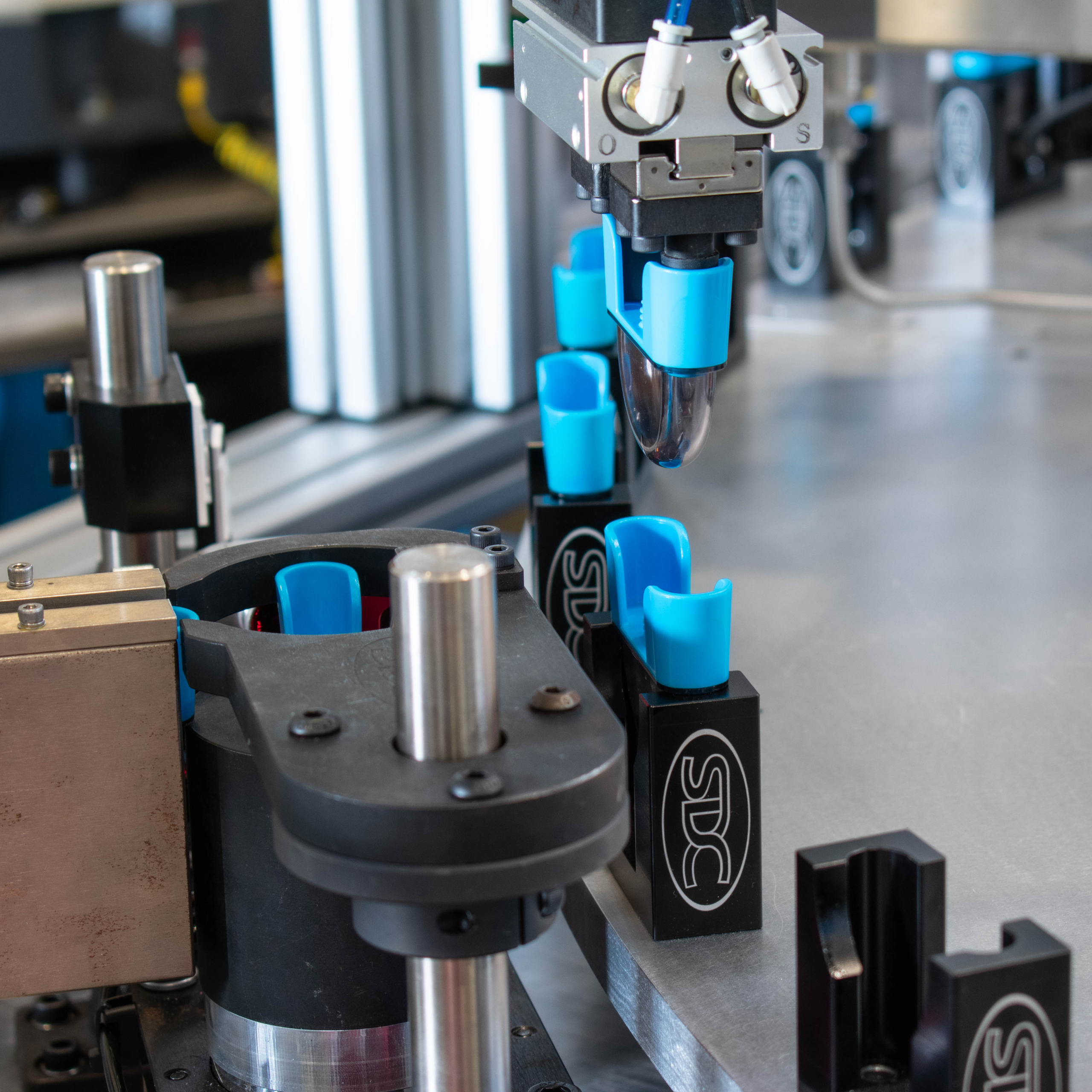 Close-up shows automated production of blocks labeled "SDC". The blocks are black, and each has a blue component being placed on it by a robotic arm.