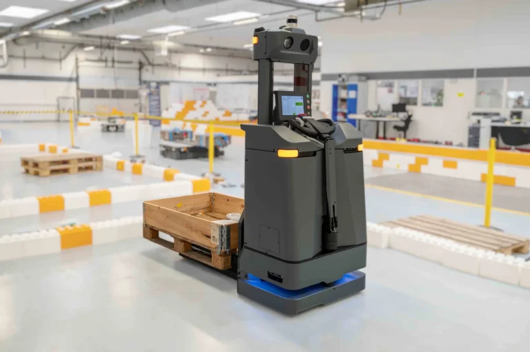 Automated guided vehicle, designed in grey, navigates a warehouse floor with color-coded lane markers, carrying a wooden pallet in an industrial setting.