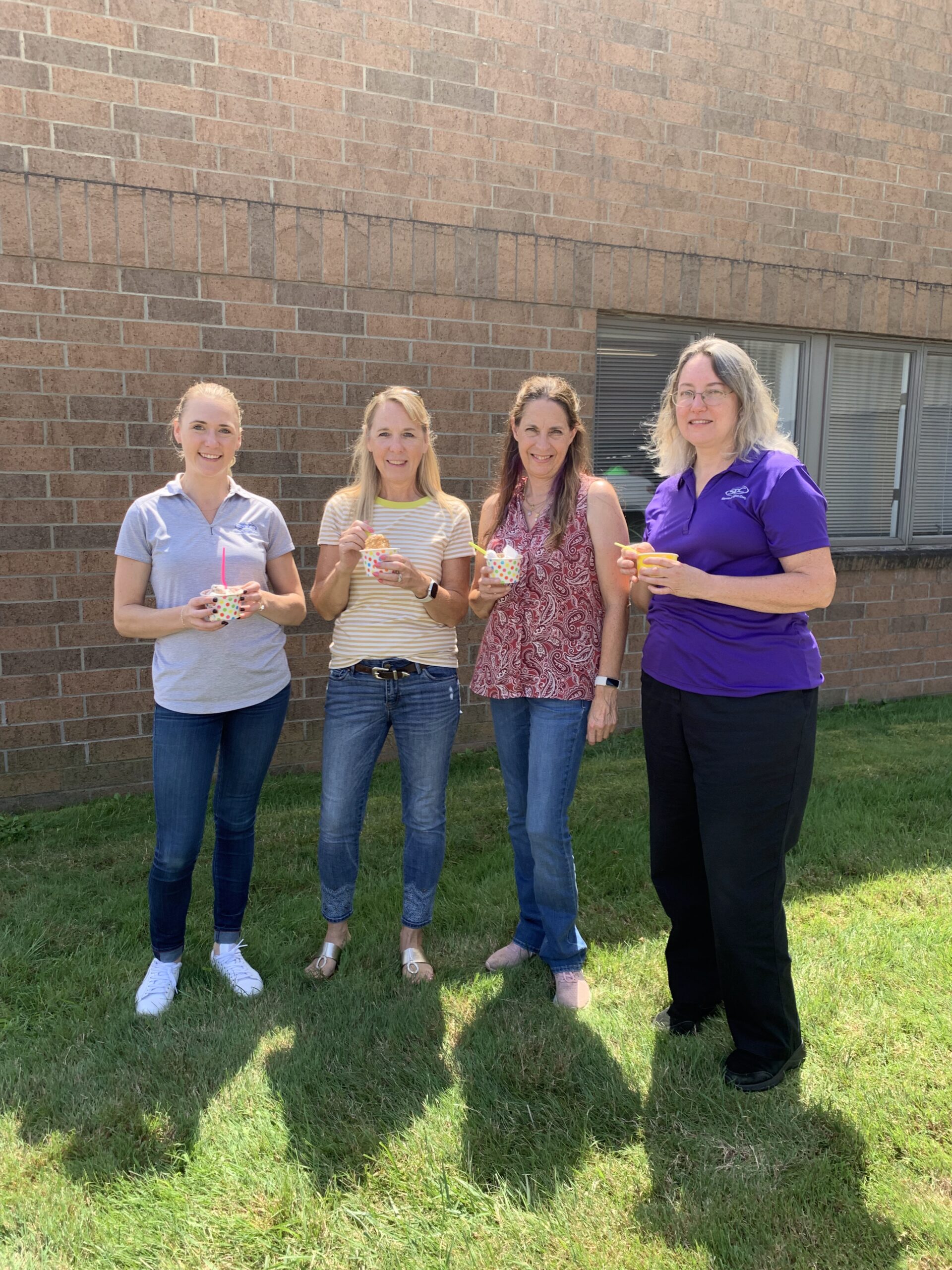 Four women stand on grass near a brick building, holding and eating ice cream in dotted cups.