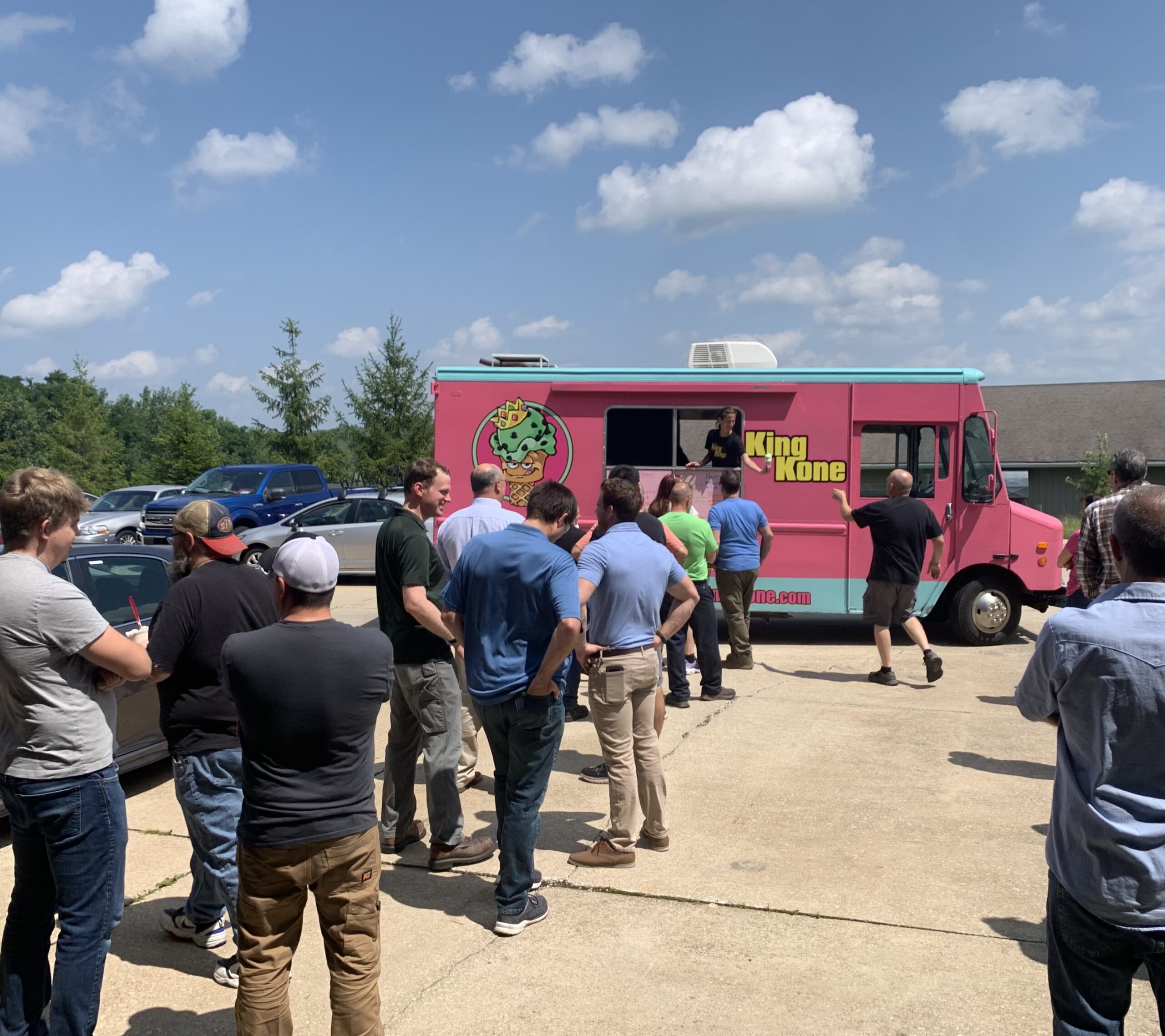 People line up at a pink "King Kone" ice cream truck on a sunny day.