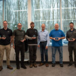 Six employees holding awards stand in front of a window. They're dressed casually and smiling.