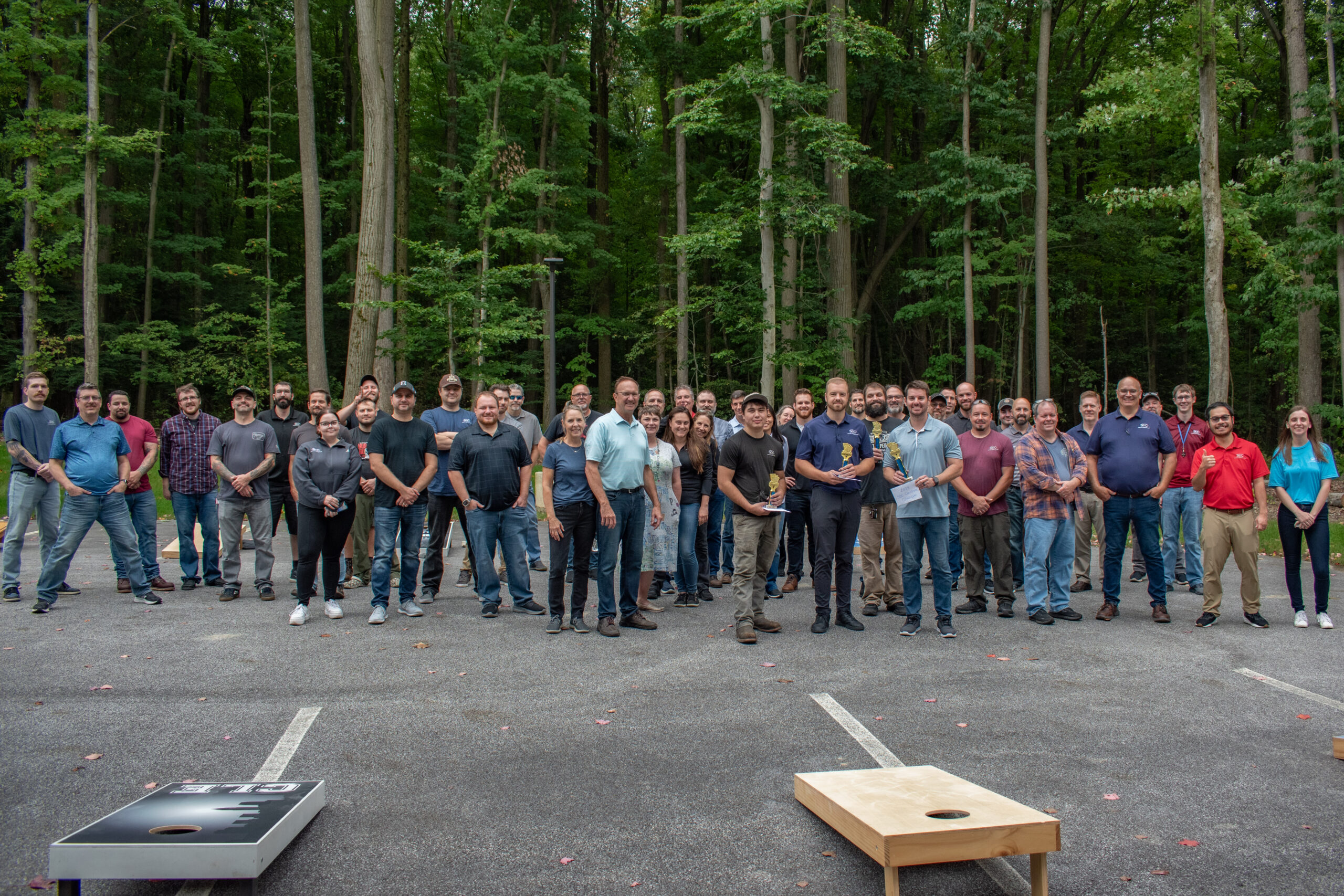 Group photo of employees outdoors after a cornhole tournament, some holding trophies. Relaxed, casual setting with trees in background.
