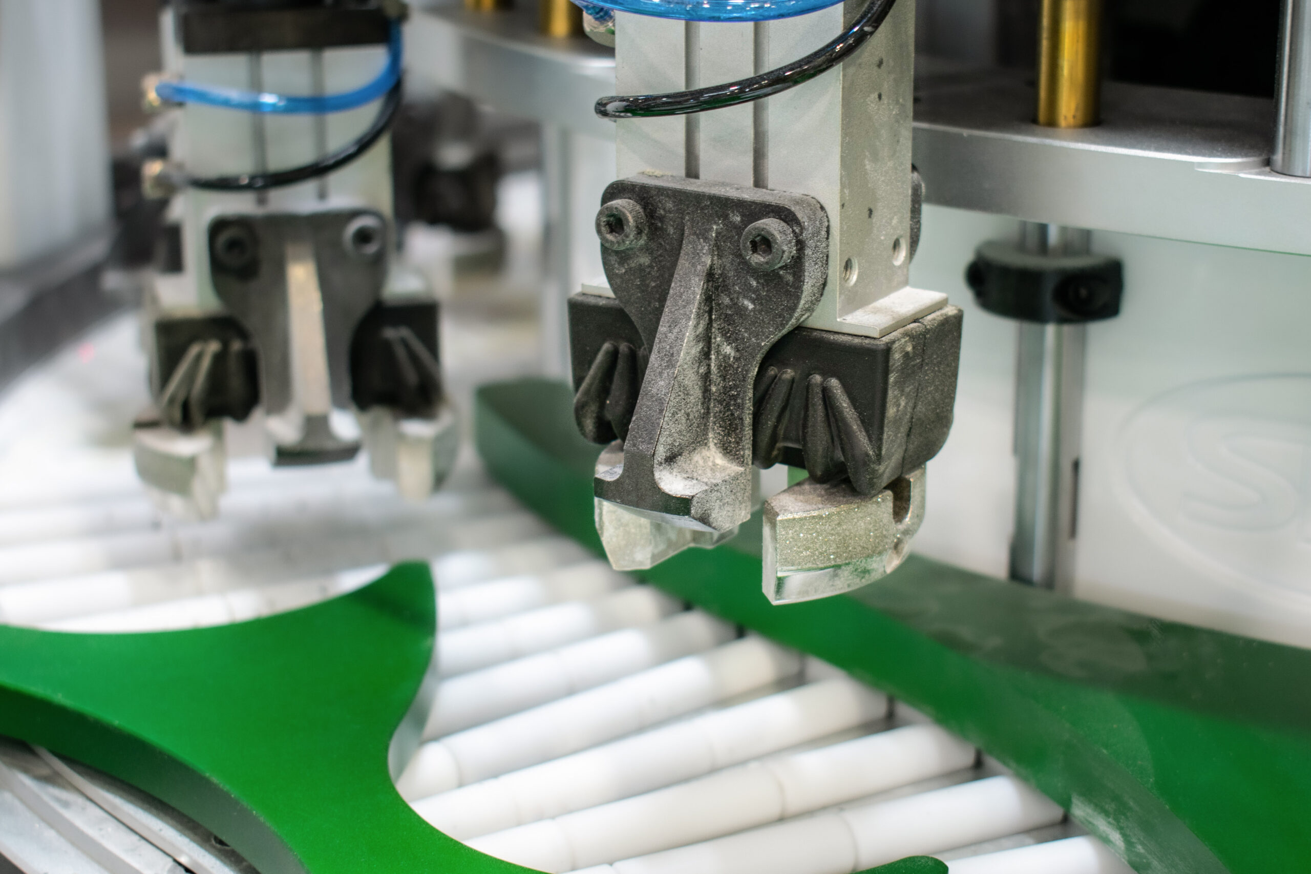 A close-up depicts an automated manufacturing process. Two robotic arms, dusty and gray, are precisely positioned above a conveyor belt with white rollers.