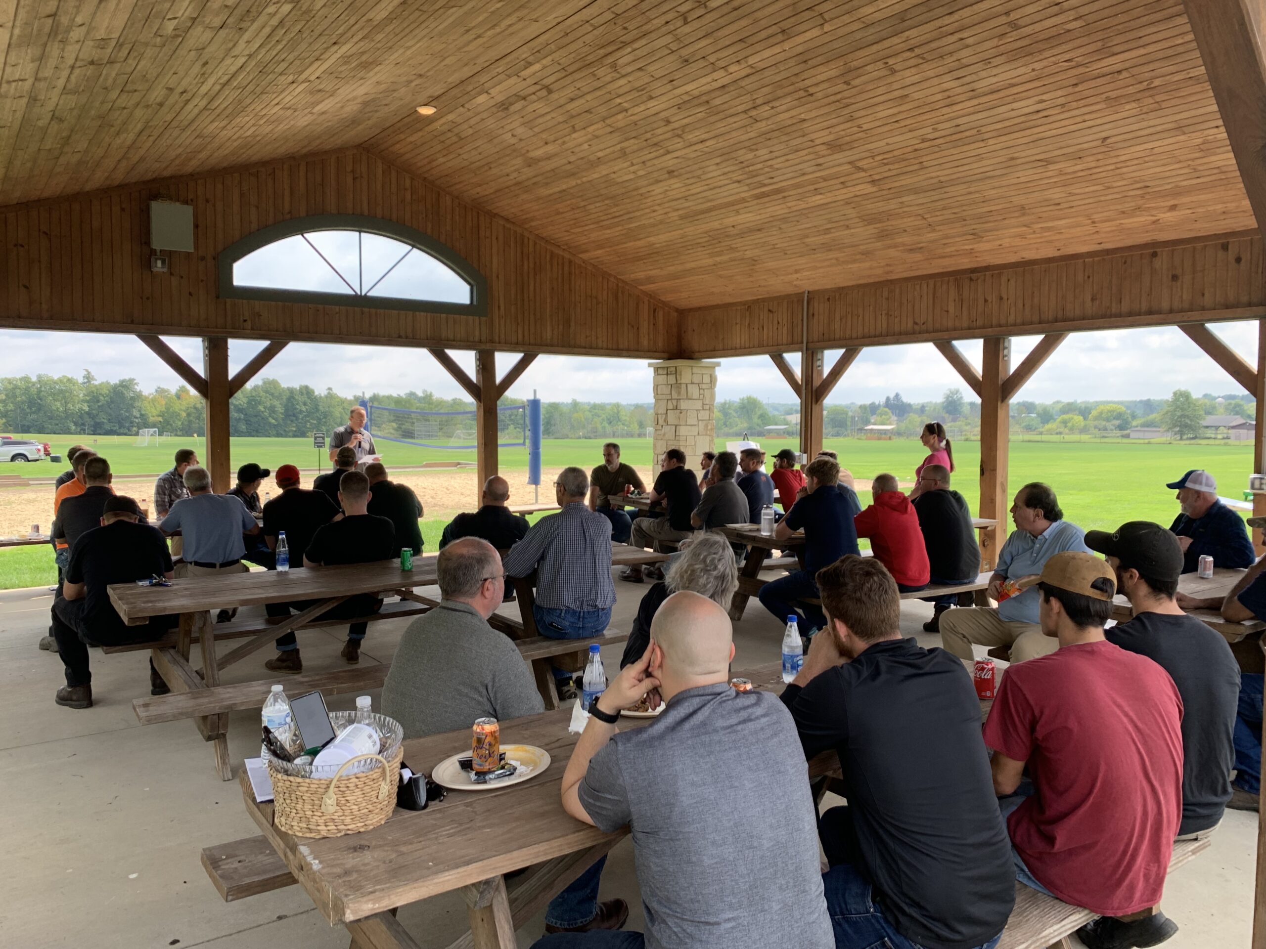 Outdoor meeting under a pavilion. A man speaks to a group seated at picnic tables.